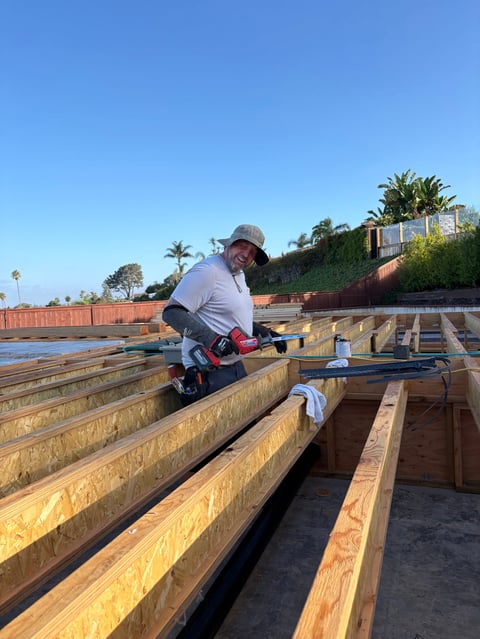 Worker in cap using power drill on wooden beam during construction on sunny day with palm trees in background