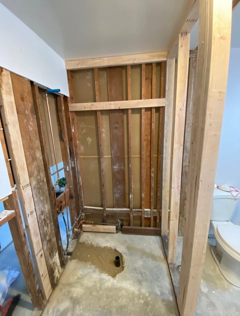 Interior of a small room under construction with exposed wooden framing, white drywall, and a concrete floor with a drainage hole