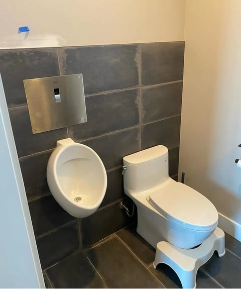 Modern bathroom with dark tile walls, featuring a white urinal on the left and a white toilet on the right with blue accents, next to a beige wall