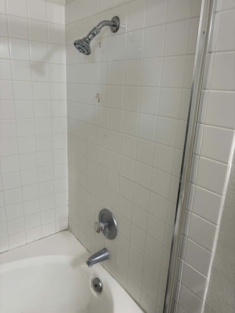 Bathroom shower with white tile walls, chrome showerhead at top, and control fixtures mounted on wall above bathtub