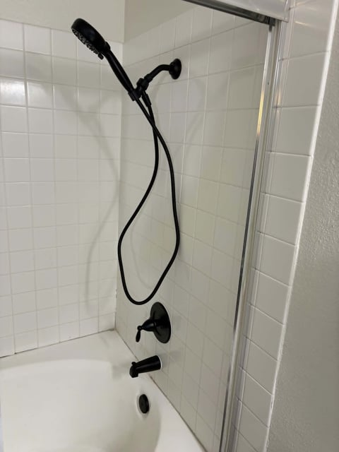 Black handheld showerhead with curved hose mounted on white tile bathroom wall above bathtub with glass shower door